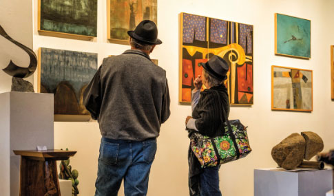 Two people wearing hats stand in an art gallery, viewing paintings on a well-lit wall. One person carries a colorful patterned bag. Sculptures and artworks are displayed around them.