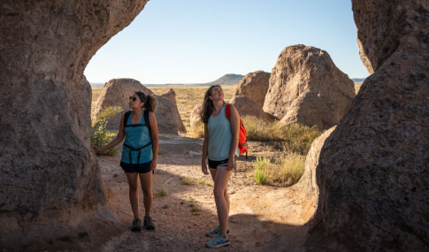 Two women with backpacks stand between large rock formations in a sunny, desert-like landscape, looking up and enjoying the scenery. Grassy plains and distant hills are visible in the background.