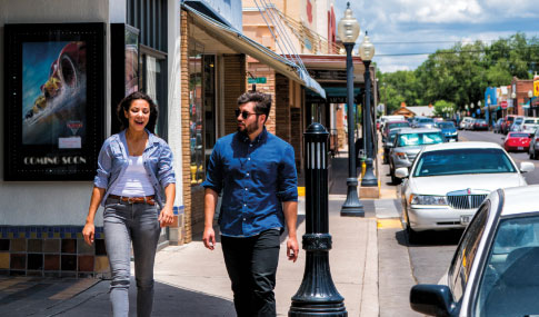 A woman and a man walk and talk on a sunny sidewalk lined with shops and parked cars. The woman is smiling and gesturing, while the man listens. A movie poster is visible on a nearby wall.