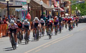 A group of cyclists in colorful jerseys race down a city street lined with shops and spectators on a sunny day. Orange barricades and street lamps are visible along the sidewalk.