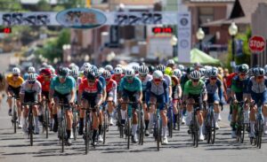 A large group of cyclists wearing colorful jerseys and helmets ride closely together on a city street during a road race, with a race banner and timer visible in the background.